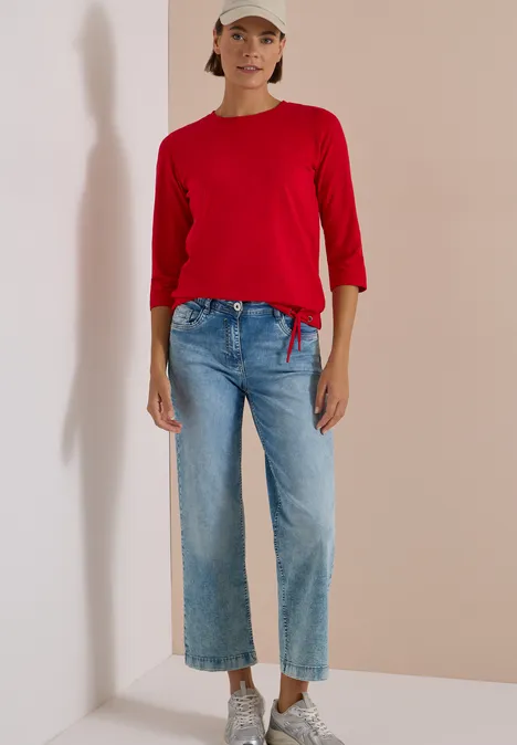 3/4-Arm Shirt mit Rundhals in Unifarbe flame red Frau in rotem Langarmshirt, weiten Jeans, weißen Sneakern und Baseballcap vor neutraler Wand.
