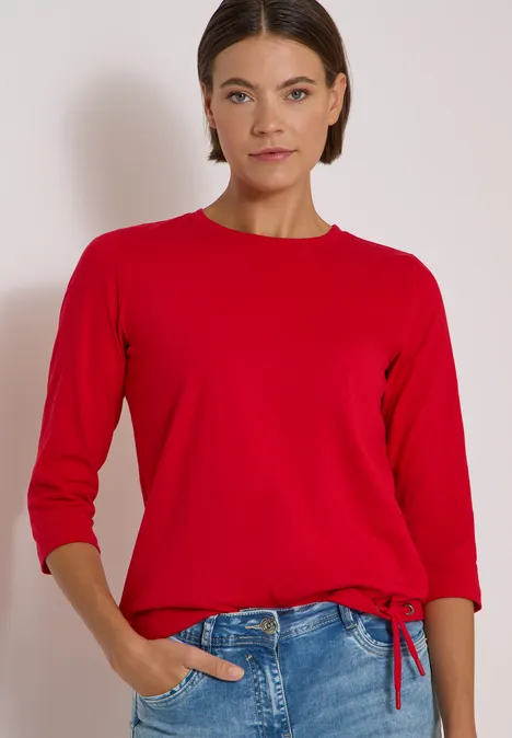 3/4-Arm Shirt mit Rundhals in Unifarbe flame red Frau vor hellem Hintergrund trägt roten Pullover mit 3/4-Ärmeln und blaue Jeans, eine Hand in Tasche.