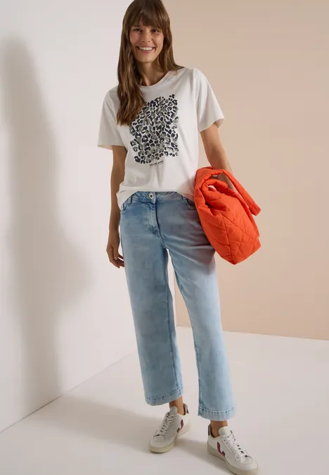 Shirt met luipaardprint en folie-details vanilla white Junge Frau lehnt an Wand, trägt weißes T-Shirt mit Print, helle Jeans, weiße Sneaker und orange Jacke.