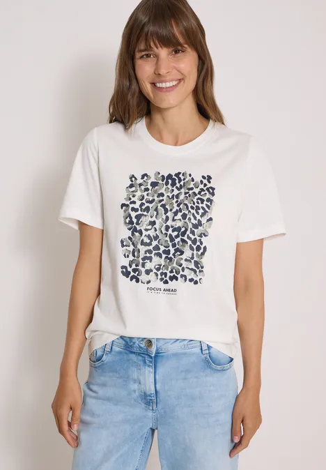 Shirt met luipaardprint en folie-details vanilla white Junge Frau in weißem T-Shirt mit abstraktem Muster und Jeans lächelt vor neutralem Hintergrund.