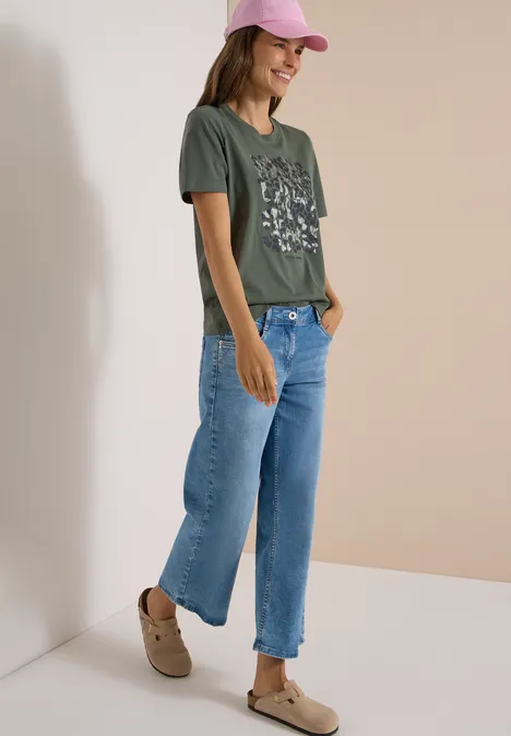 Shirt met luipaardprint en folie-details dried olive green Frau in grünem Print-Shirt, weiten cropped Jeans, beigen Schuhen und rosa Cap lehnt lächelnd an Wand.
