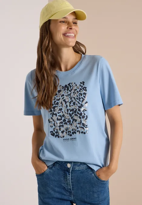 Shirt met luipaardprint en folie-details easy blue Frau mit hellblauem T-Shirt mit Leopardenmuster-Herz, blauer Jeans und gelber Cap, lächelnd vor neutralem Hintergrund.