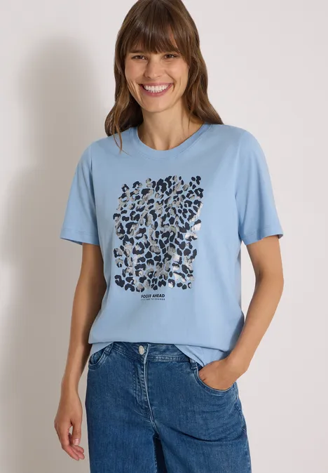 Shirt met luipaardprint en folie-details easy blue Frau lächelt, trägt hellblaues T-Shirt mit abstraktem Animal-Print und blaue Jeans, eine Hand in der Tasche.