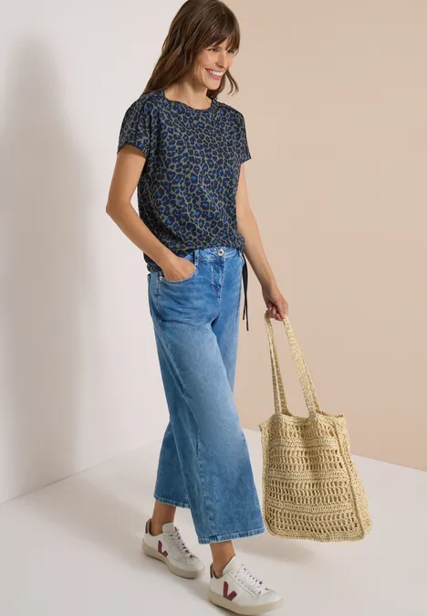Frau in Leopardenmuster-Shirt, weiten Jeans und Basttasche lächelnd