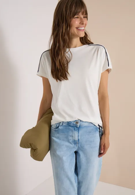 Frau in weißem T-Shirt und hellblauen Jeans hält ein grünes Kissen und lächelt.
