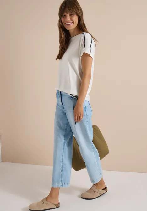 Junge Frau in weißem T-Shirt, hellblauen Jeans und Loafers, die lächelnd eine Tasche hält.
