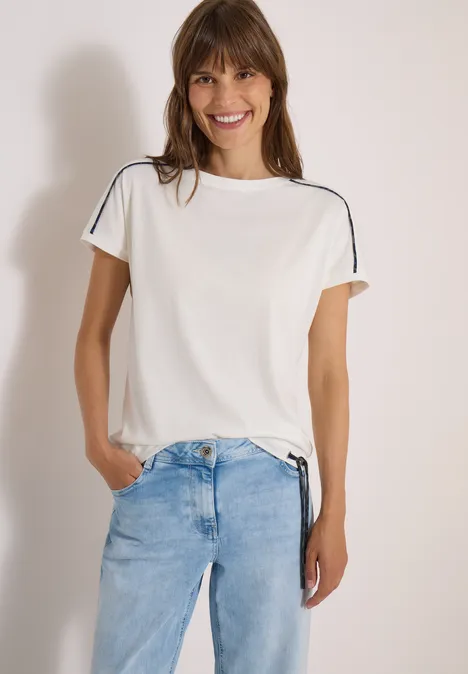 Eine lächelnde Frau trägt ein weißes T-Shirt mit Schulterstreifen und helle Jeans und hat die Hände in den Taschen.