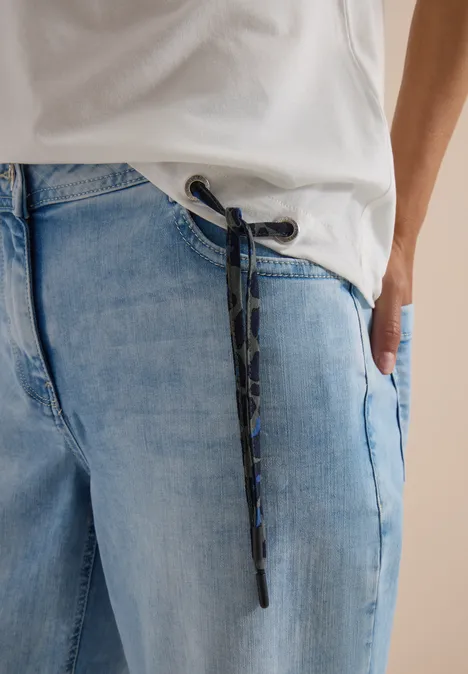 Person trägt hellblaue Jeans mit Camouflage-Kordel, Hand in Tasche und weißem T-Shirt.