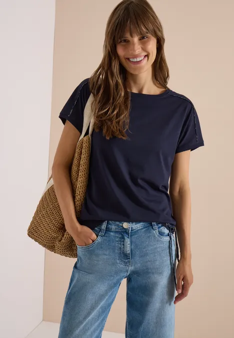 Junge Frau in casual Outfit: dunkelblaues T-Shirt, Jeans und Basttasche, lächelnd vor neutralem Hintergrund.