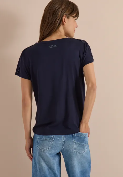 Rückenansicht einer Frau in dunkelblauem T-Shirt und Jeans vor neutralem Hintergrund.