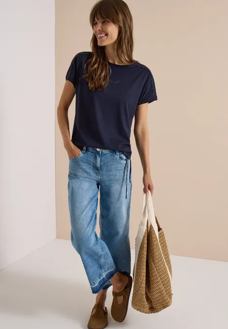 Frau in dunkelblauem T-Shirt, weiten Jeans und braunen Mokassins trägt eine große geflochtene Tasche.