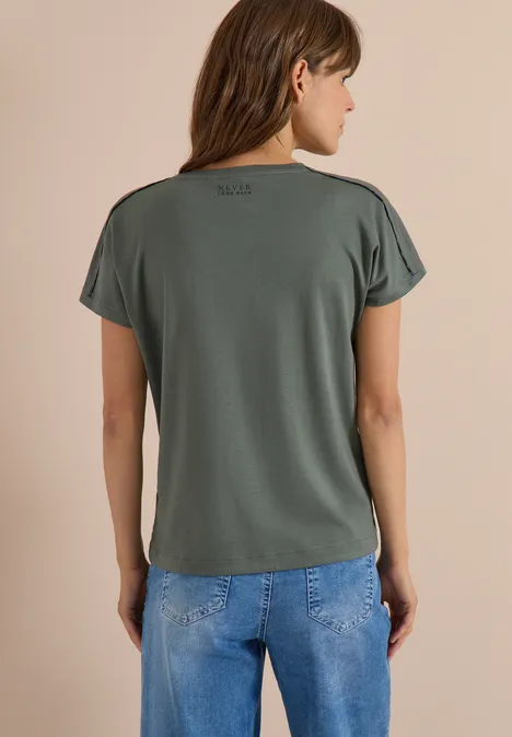 Frau steht im Profil, trägt grünes kurzärmeliges Shirt und blaue Jeans vor neutralem Hintergrund.