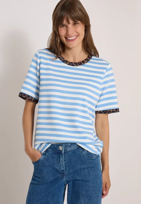Eine lächelnde Frau in hellblau-weiß gestreiftem T-Shirt mit dekorativem Kragen und Jeans vor neutralem Hintergrund.