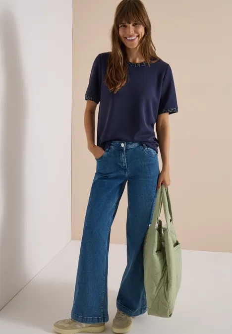 T-shirt à col rond avec des détails léopard urban dark blue Eine lächelnde Frau trägt ein dunkelblaues T-Shirt, weite Jeans und hält eine grüne Tasche.