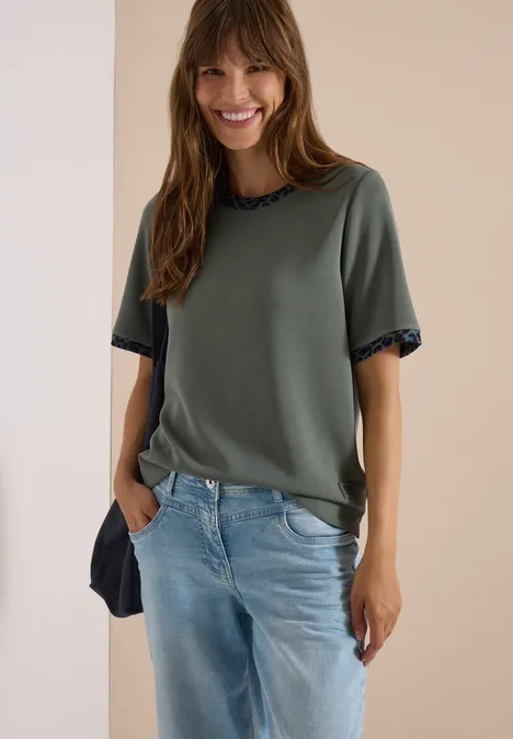 Eine lächelnde Frau trägt ein grünes Shirt und blaue Jeans.