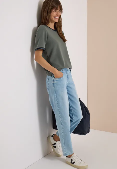Junge Frau mit grünem Shirt, hellblauen Jeans und Sneakern lehnt an Wand, lächelt und trägt Tasche.
