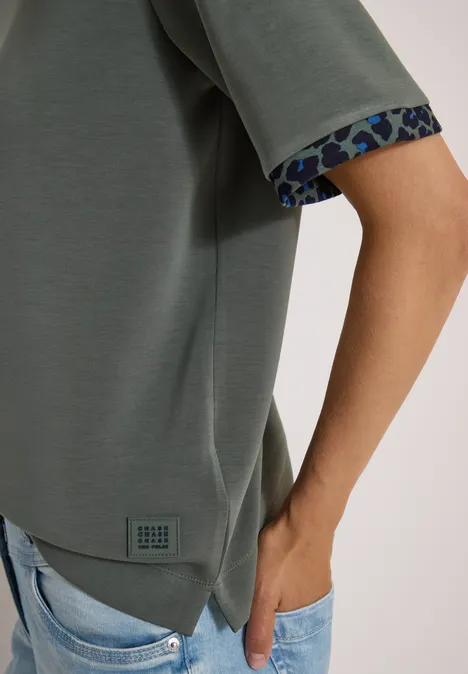 Olivgrünes Shirt mit gemusterter Ärmelkante und hellblaue Jeans, Hand in Tasche.