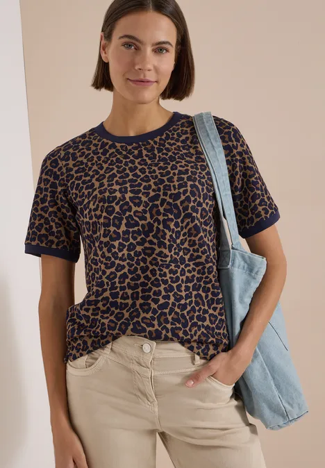 Frau trägt kurzärmeliges Leopardenmuster-Shirt, helle Hose und blaue Tasche, lächelt vor neutralem Hintergrund.