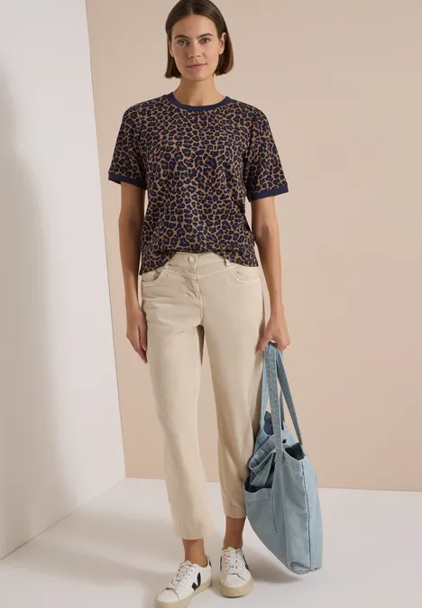 Eine Frau trägt ein T-Shirt mit Leopardenmuster, eine helle Chinohose, weiße Sneaker und hält eine blaue Stofftasche.
