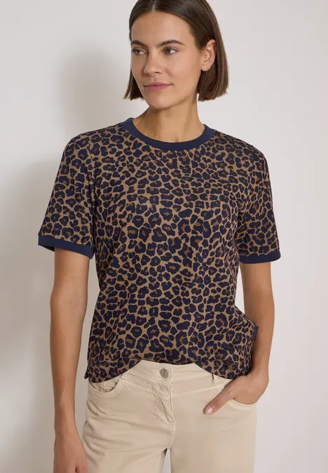 Frau in kurzarm Leo-Shirt (blau-braun) und hellen Hosen vor schlichtem Hintergrund.