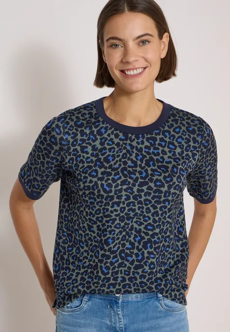 Frau mit kurzärmeligem Leo-Pullover in Blau-Grau, lächelnd vor hellem Hintergrund.