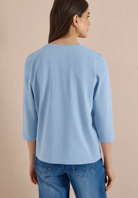 Koszulka z rękawami 3/4 z napisem w stylu lampartowym easy blue Kobieta widziana od tyłu w luźnej jasnoniebieskiej bluzce 3/4 rękaw i dżinsach na neutralnym tle