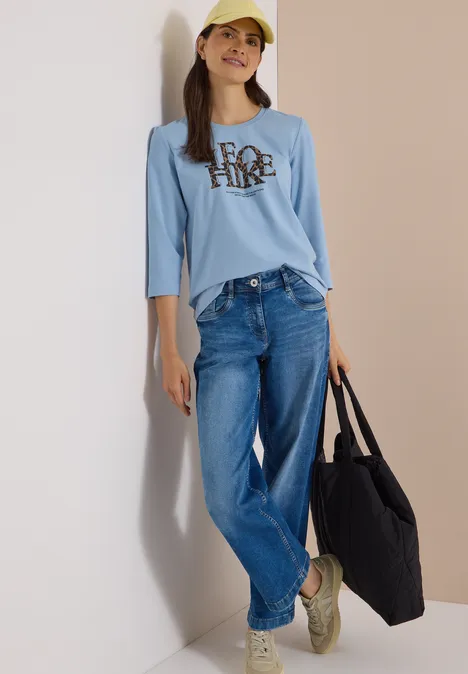 Koszulka z rękawami 3/4 z napisem w stylu lampartowym easy blue Kobieta w żółtej czapce, jasnoniebieskiej bluzce z długim rękawem, dżinsach i czarnej torbie.
