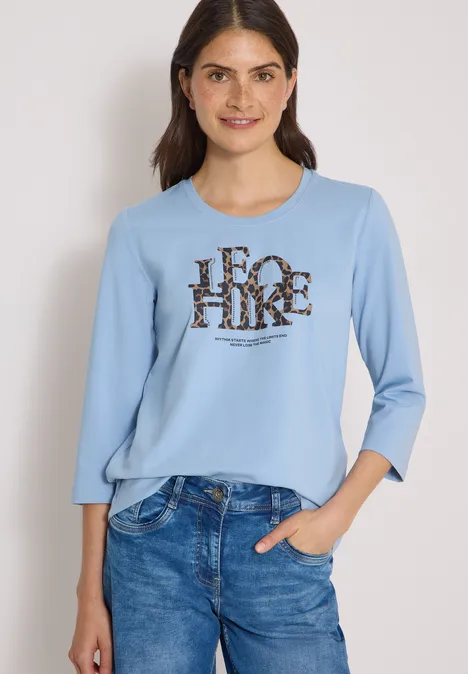Koszulka z rękawami 3/4 z napisem w stylu lampartowym easy blue Uśmiechnięta kobieta w niebieskim swetrze z napisem 'FOR HIKE' i dżinsach na neutralnym tle