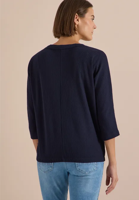 Frau in marineblauem 3/4-Arm-Shirt und Jeans, Rückansicht vor neutralem Hintergrund.