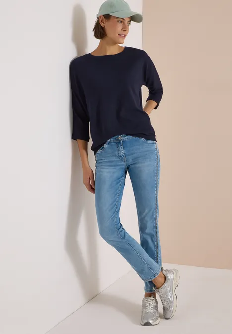 Frau mit Cap, dunkelblauem 3/4-Shirt, hellen Jeans und Sneakers lehnt entspannt an einer Wand.