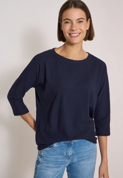 Eine Frau mit kurzem braunem Haar trägt ein dunkelblaues Shirt und Jeans, sie lächelt.