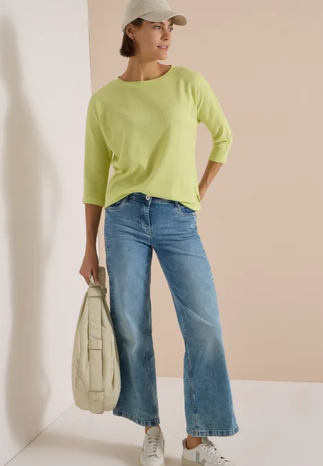 Eine Frau lehnt an der Wand, trägt hellgrünen Pullover, Jeans, Cap und Tasche.