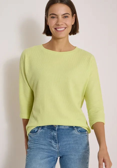 Eine lächelnde Frau trägt einen hellgrünen Pullover mit 3/4-Ärmeln und blaue Jeans vor neutralem Hintergrund.