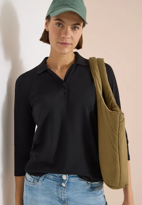 Frau trägt schwarzen Poloshirt, grüne Mütze und olivgrüne, gesteppte Umhängetasche