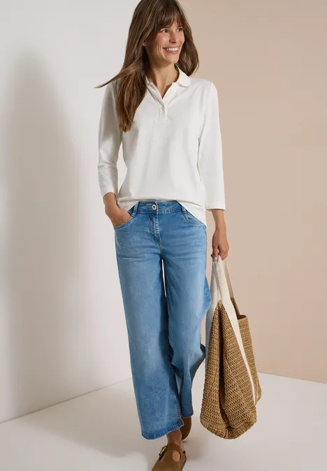 Frau in weißer Bluse und weitem Jeans, lächelnd vor zweifarbiger Wand, hält Korbtasche.