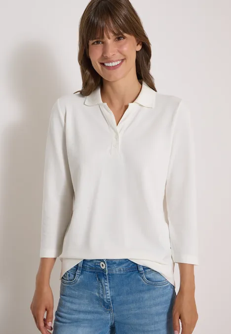 Frau in weißem Langarm-Poloshirt und Jeans, lächelt vor neutralem Hintergrund.