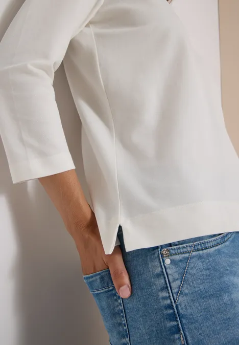 Frau trägt weißes Shirt mit 3/4-Ärmeln und blaue Jeans, Hand in Hosentasche.