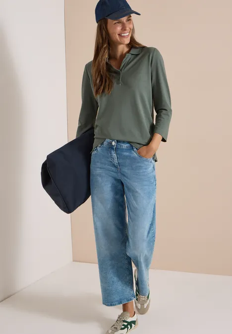 Frau mit blauer Jeans, olivgrünem Shirt und Cap hält navy Jacke, lässiger Alltagslook.