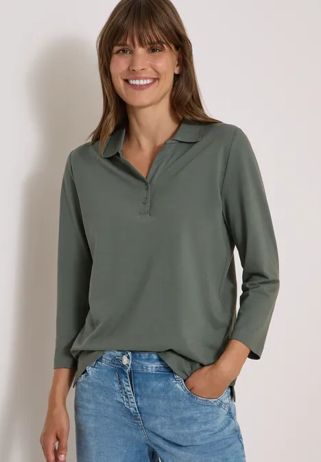 Frau mit langärmligem grünen Poloshirt, Jeans, Hände in den Taschen, lächelnd vor weißem Hintergrund.
