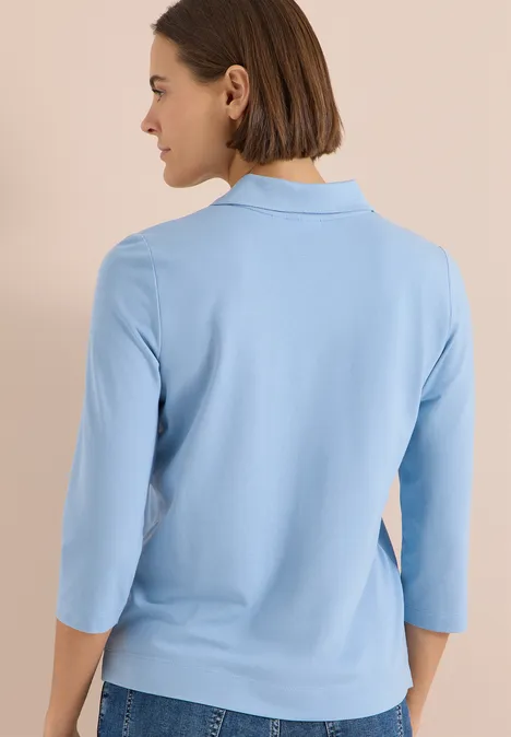 Frau mit dunklem Bob trägt hellblaues 3/4-Arm-Shirt, steht blickabgewandt vor neutralem Hintergrund.