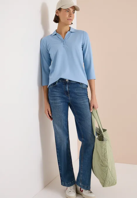 Frau trägt hellblaues Poloshirt, weite Jeans und hält eine grüne Stofftasche vor zweifarbigem Hintergrund.