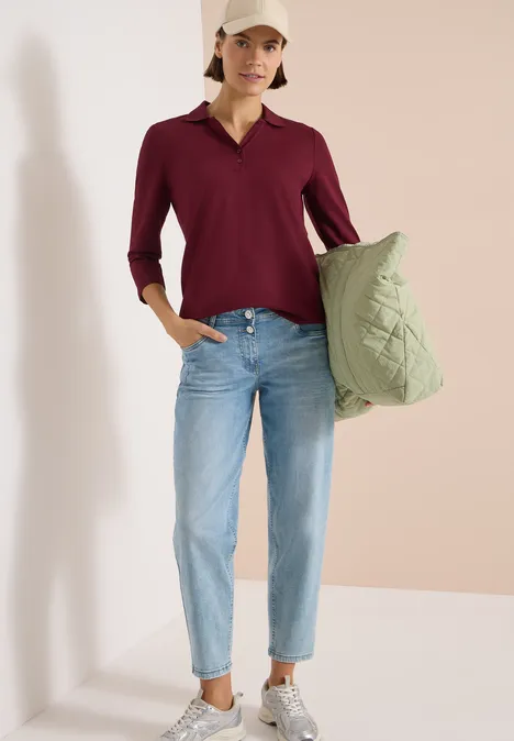 Frau trägt eine grüne Stepp-Tasche, bordeauxrote Bluse und helle Jeans vor hellem Hintergrund.