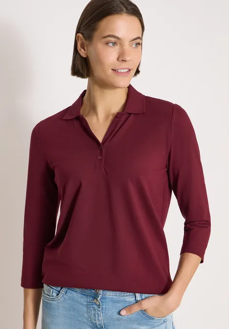 Eine Frau trägt ein burgunderfarbenes Langarmshirt und Jeans, steht vor neutralem Hintergrund.