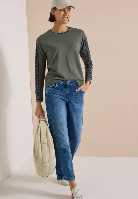 Frau mit grüner Langarmbluse, gemusterten Ärmeln, blauen Jeans, Sneakern und Tasche lehnt lässig an einer Wand.