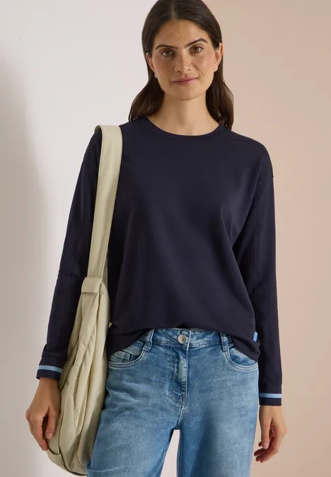 Vrouw in marineblauwe trui en jeans met beige schoudertas, voor een neutrale achtergrond.