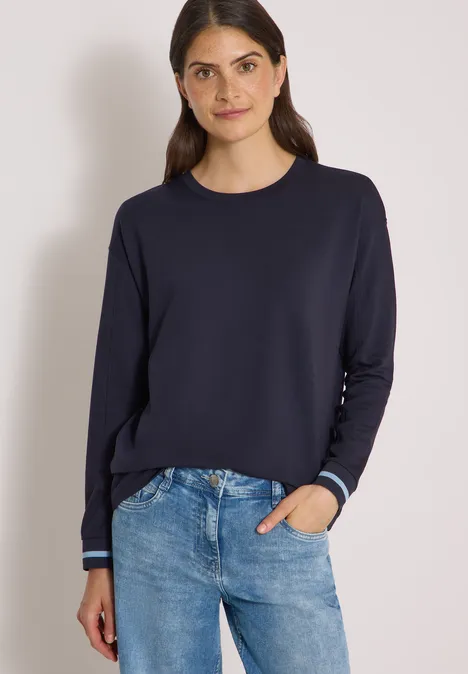 Frau in dunkelblauem Langarmshirt und Jeans vor hellem Hintergrund