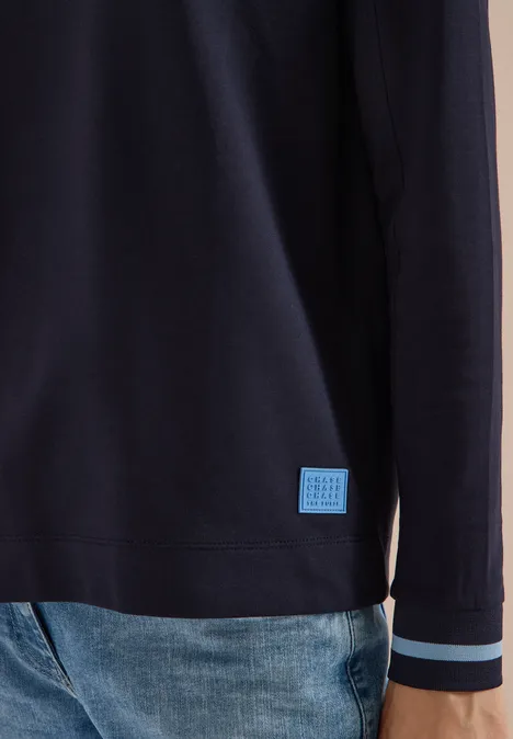 Dunkelblauer Pullover mit hellen Streifen an den Bündchen und kleinem Logo-Patch am Saum, kombiniert mit Jeans.