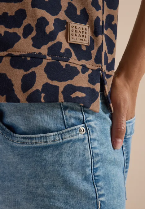 Nahaufnahme einer Person in hellblauen Jeans, Hand in Tasche, trägt ein Oberteil mit Leopardenmuster und Label.