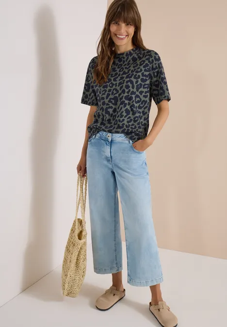 Frau in Leoparden-T-Shirt und hellen Jeans-Culottes, trägt Korbtasche und bequeme Slipper.