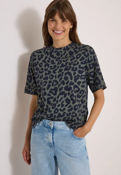 Frau in grünem Leo-T-Shirt und hellblauen Jeans, lächelt mit einer Hand in der Tasche.
